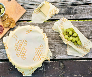 Küche ohne Plastik. Auf dem Bild sind Trauben, ein Stück Käse und eine Schale mit drei verschiedenen, von der Größe her, Bienenwachstücher eingewickelt.  Dabei ein Holzbrett mit Brottscheiben.