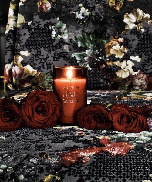 Bio Duftkerze mit Vanille Duft in einem feuerfesten roten Glas mit Korkdeckel und die Beschriftung "All of me loves all of you" umgebend von Rosen und Blumen.
